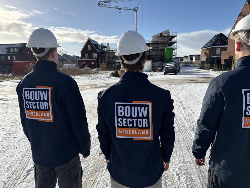 Drie vakmensen van Bouwsector Nederland in blauwe jassen met een hijskraan boven de bouwplaats